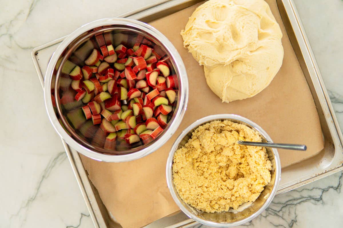 sliced rhubarb in a bowl and streusel topping in a bowl and yeast dough on a baking sheet