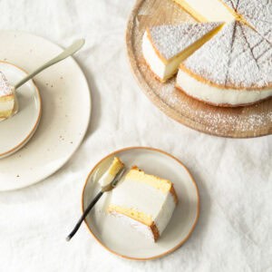Käse-Sahne-Torte slice on a plate and the rest of the cake on a wooden cakestand behind it.