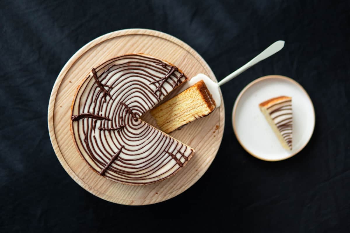 Top view of a Baumkuchen on a wooden cake stand with a slice of it sitting on a plate next to it.