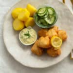 Beer-battered cod topped with lemon sitting next to remoulade sauce, potatoes and cucumber salad on a white plate.