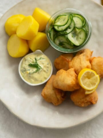 Beer-battered cod topped with lemon sitting next to remoulade sauce, potatoes and cucumber salad on a white plate.