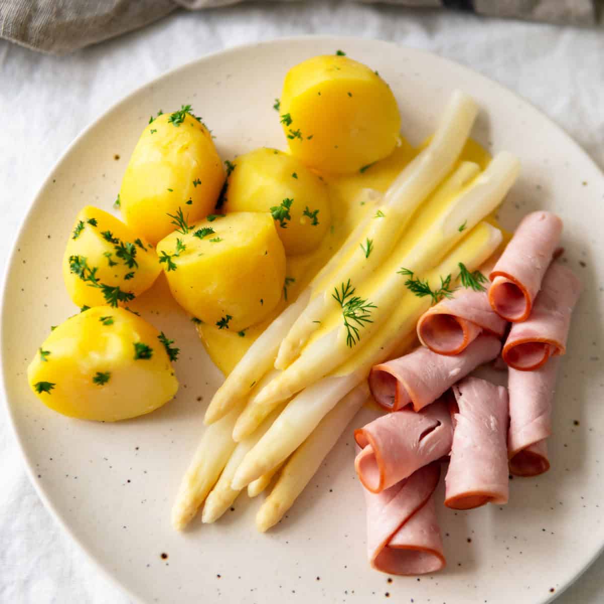 White asparagus on a plate with easy hollandaise sauce, boiled potatoes and rolled up ham slices.