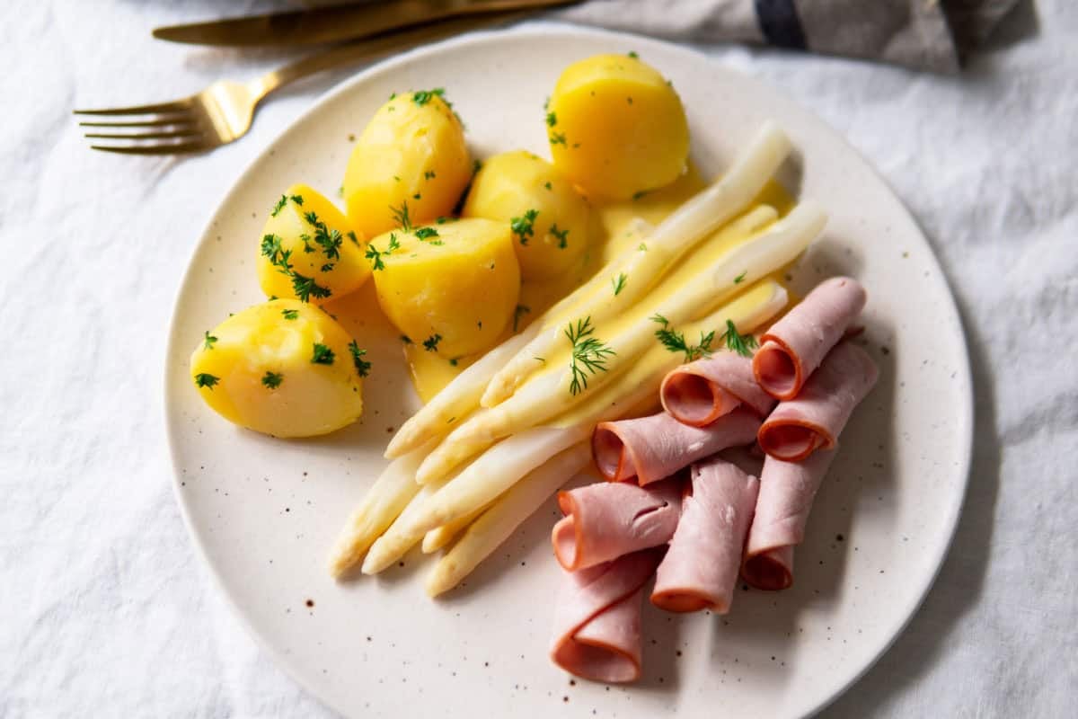 White asparagus on a plate with easy hollandaise sauce, boiled potatoes and rolled up ham slices.