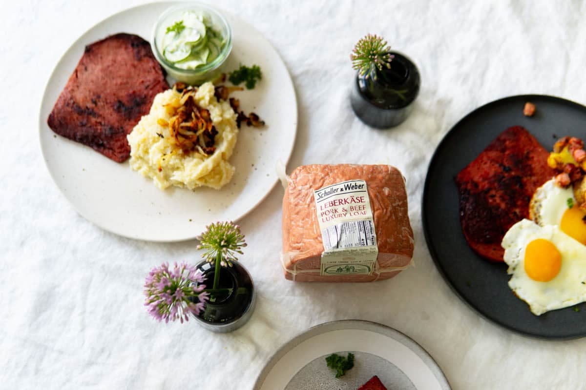 A packaged loaf of Leberkäse with cooked versions on plates around it.