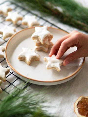 A child holding a star shaped cinnamon cookie called Zimtsterne next to a cooling rack full of Zimtsterne.