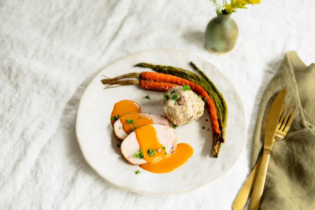 Sliced pork loin on a plate with sauce, a bread dumpling and roasted veggies.