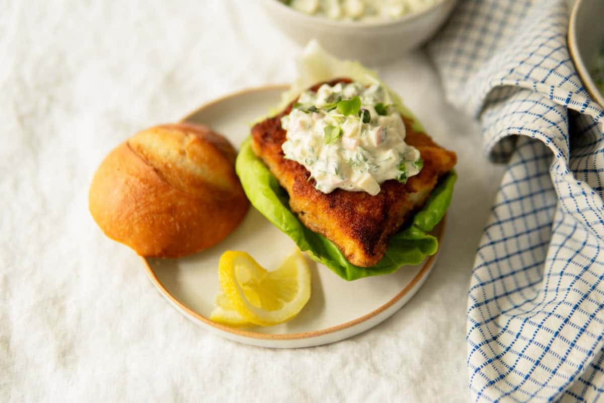 Fried fish sandwich on a plate topped with remoulade sauce, with top bun laying off to the side of the plate.