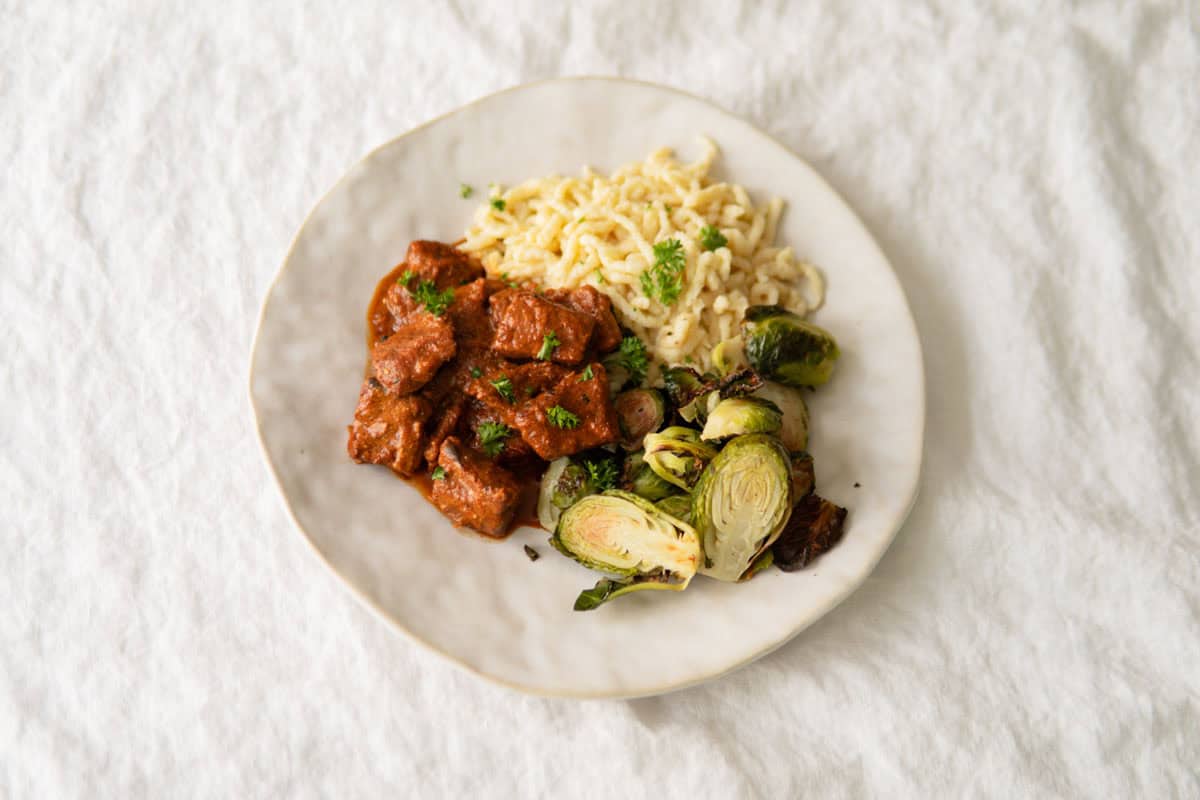 German pork goulash on a plate with spätzle noodles and brussel sprouts.